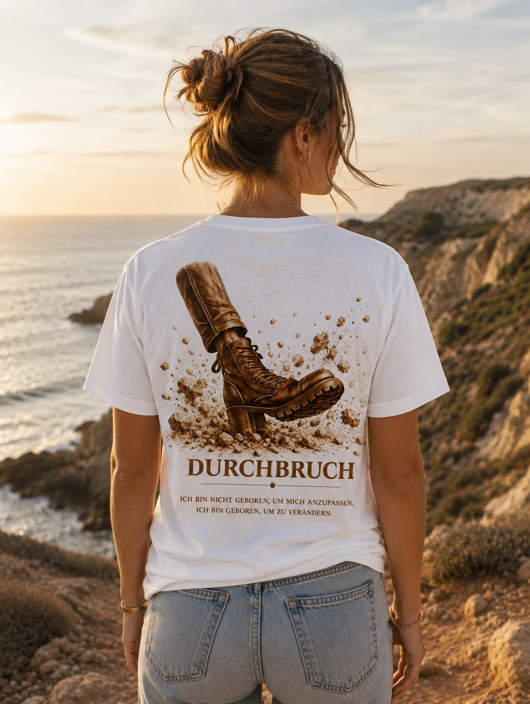 Getragen: Frau von hinten im weißen Durchbruch-Shirt an einer Küste bei Sonnenuntergang, Rückenmotiv Stiefel bricht den Boden auf, Schriftzug DURCHBRUCH